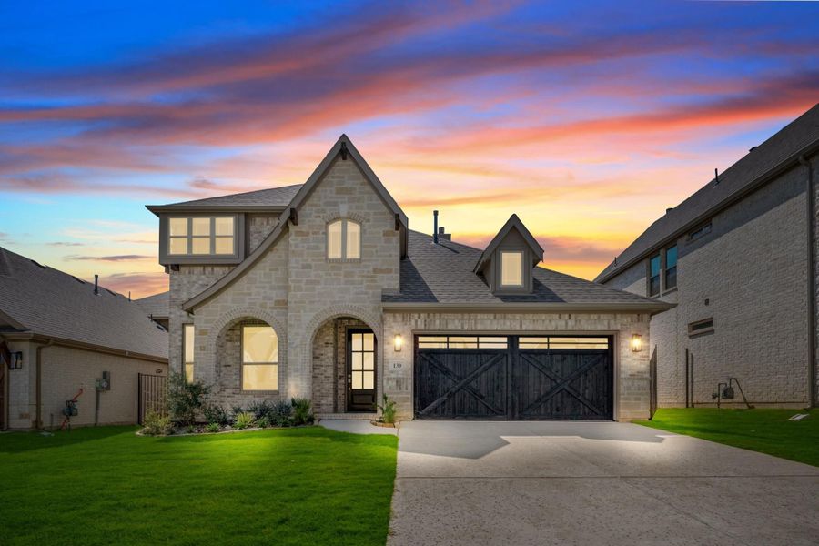 Two-story stone home with arched entry, dark barn-style garage door, and green front lawn at dusk