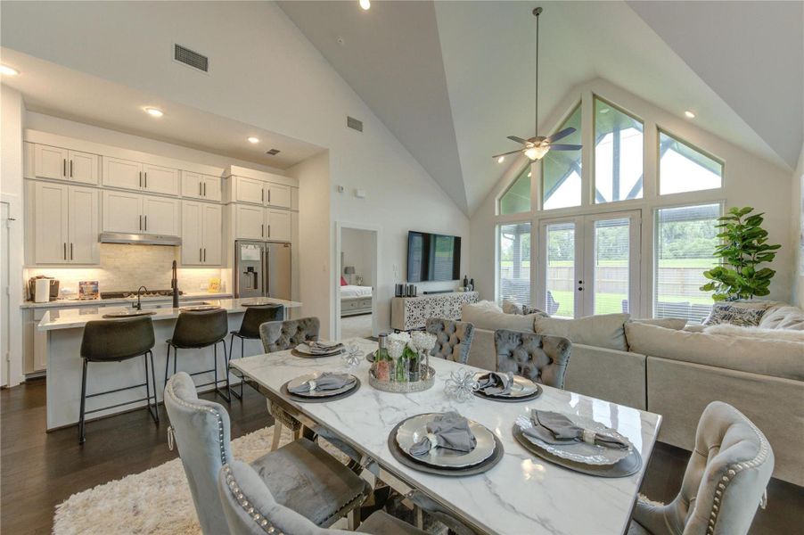 This photo showcases an elegant open-concept living space with a vaulted ceiling, featuring a modern kitchen with a large island, a cozy dining area, and a spacious living room. It has large windows providing ample natural light and a view of the backyard, creating a bright and inviting atmosphere.