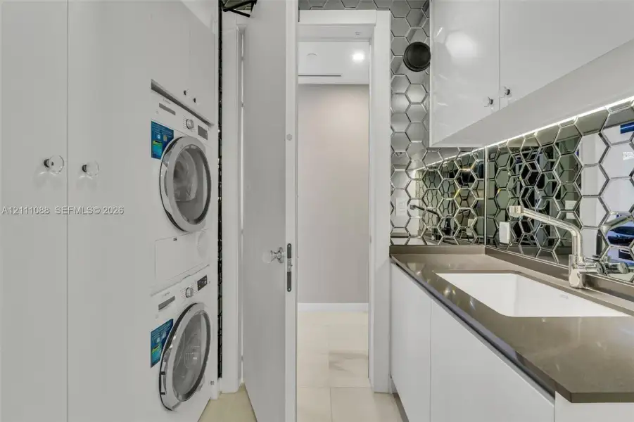 Utility space featuring high-gloss white cabinetry, integrated stacking laundry appliances, stone-finish countertops with an under-mount sink, and a metallic hexagonal tile backsplash Utility space featuring high-gloss white cabinetry, integrated stacking laundry appliances, stone-finish countertops with an under-mount sink, and a metallic hexagonal tile backsplash