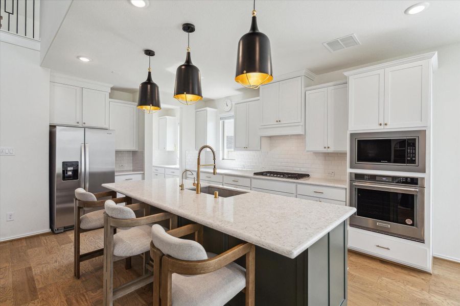 The stunning kitchen is a true showpiece, featuring upgraded appliances, white cabinetry with a classic subway tile backsplash, and a striking island painted in a contrasting dark tone with an ivory-hued countertop. Black pendant lights with warm brass interiors add an elegant focal point, balancing modern style with timeless detail.