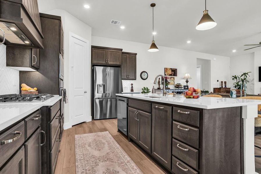 Kitchen featuring a substantial island with breakfast bar, undermount sink with gooseneck brushed nickel faucet, wood-finish flooring, dark wood cabinetry, a white herringbone tile backsplash, walk-in pantry and stainless steel appliances including a propane gas cooktop, electric built-in oven and microwave open to living and breakfast rooms