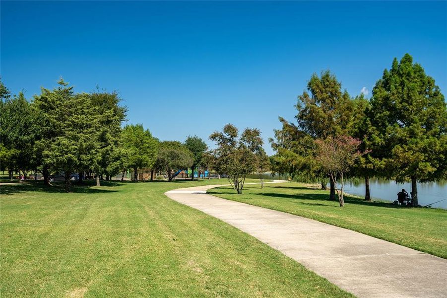 Paved pathway winding through a grassy parkland, bordered by mature trees and a tranquil body of water Paved pathway winding through a grassy parkland, bordered by mature trees and a tranquil body of water