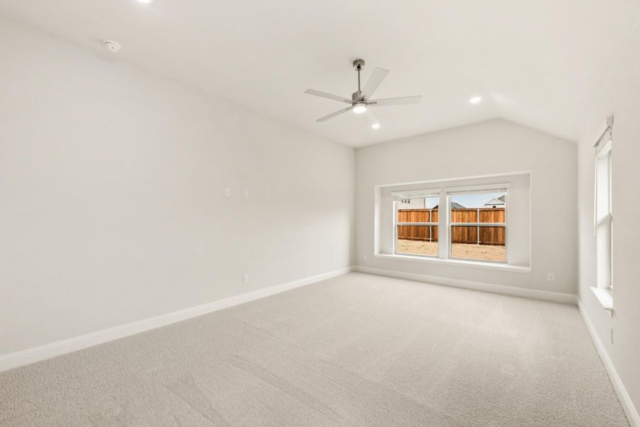 Empty bedroom with vaulted ceiling, ceiling fan, carpet flooring, and large window overlooking backyard fence Empty bedroom with vaulted ceiling, ceiling fan, carpet flooring, and large window overlooking backyard fence