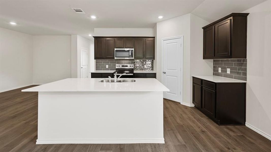 Modern kitchen featuring a large white island with an integrated sink, dark wood cabinetry, white countertops, a subway tile backsplash, and stainless steel appliances Modern kitchen featuring a large white island with an integrated sink, dark wood cabinetry, white countertops, a subway tile backsplash, and stainless steel appliances