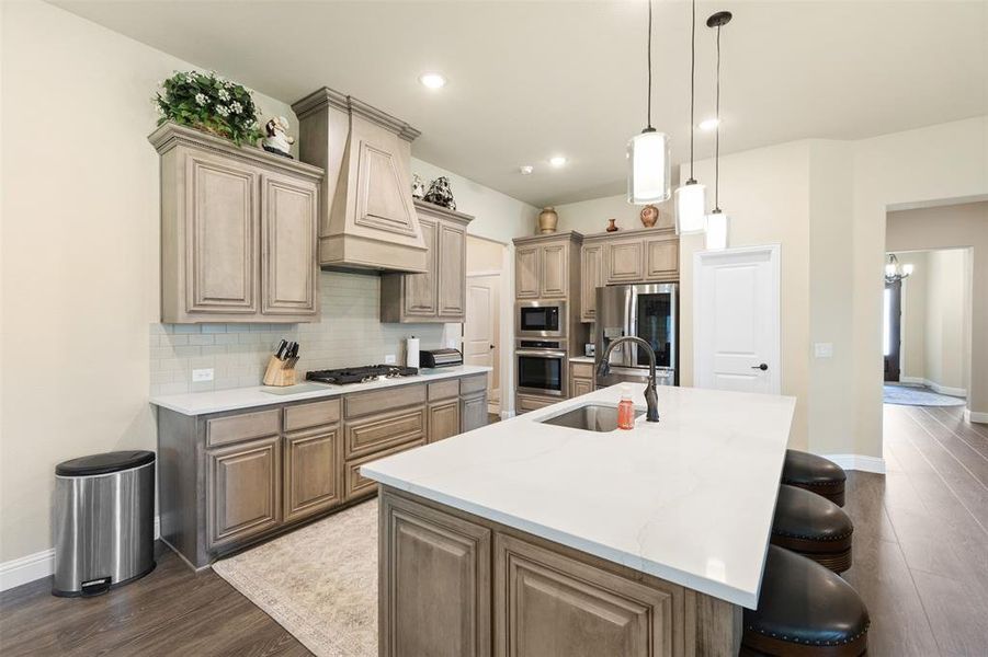 Kitchen featuring an expansive center island with a built-in sink, pendant lighting, wood-finish flooring, light-toned cabinetry, and stainless steel appliances Kitchen featuring an expansive center island with a built-in sink, pendant lighting, wood-finish flooring, light-toned cabinetry, and stainless steel appliances