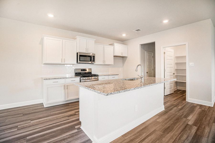 This kitchen features modern white cabinets, granite countertops, and stainless steel appliances. It has a spacious open layout with luxury vinyl plank flooring and an adjacent pantry for extra storage.