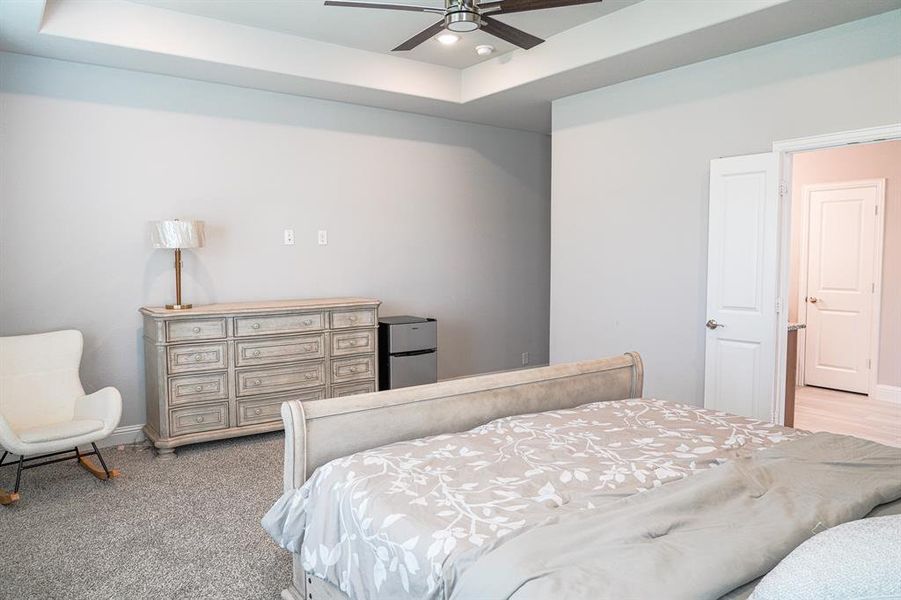 Bedroom featuring freestanding refrigerator, a tray ceiling, a ceiling fan, and light carpet