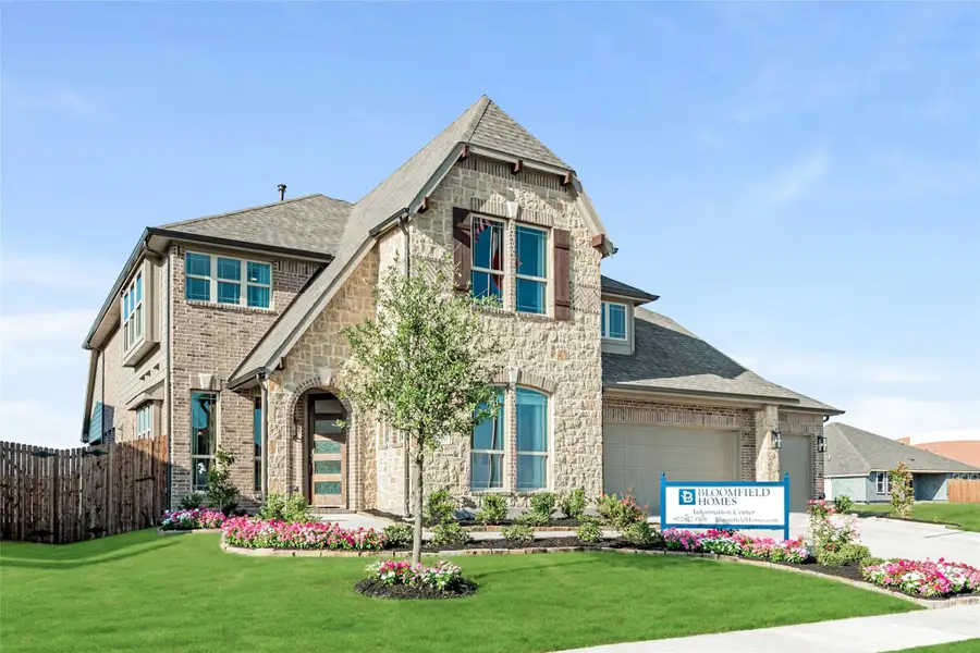 Two-story Bloomfield Homes model with brick and stone exterior, two-car garage, and landscaped front yard with flowers.