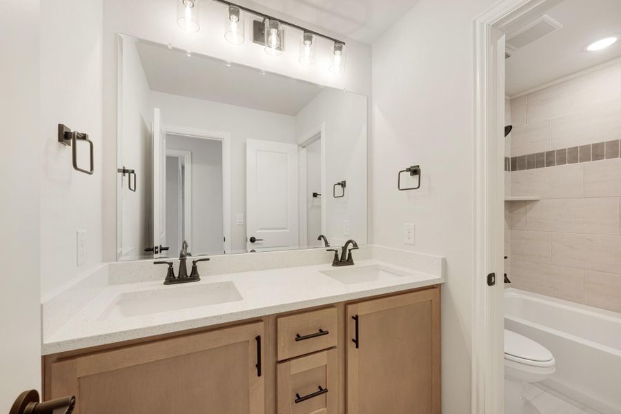 Bathroom with double sink vanity, light wood cabinets, white countertop, and tiled tub-shower combo with built-in shelves