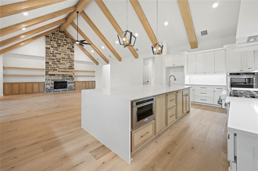 Open-concept kitchen and living area featuring vaulted ceilings with exposed wood beams, a stone-front fireplace, built-in shelving, a large central island with an integrated microwave, and stainless steel appliances