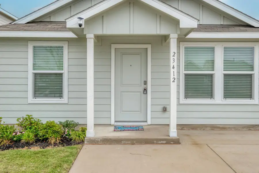 Light-colored siding exterior featuring a covered entry, two front windows with blinds, a concrete walkway, and landscaped garden beds