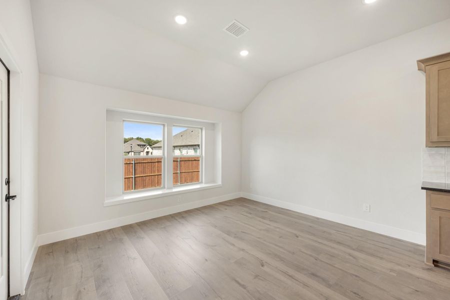 Empty dining area with vaulted ceiling, light wood floors, and a double window overlooking a backyard fence.