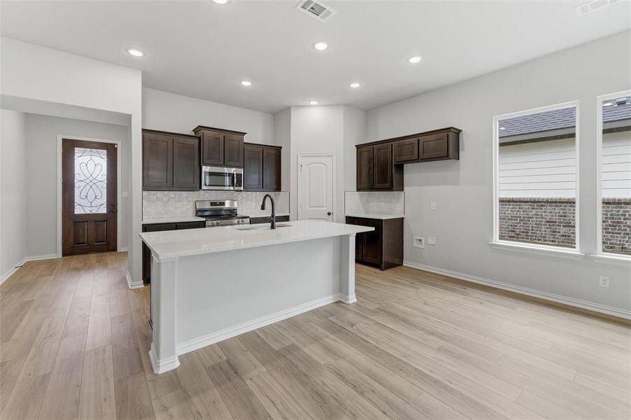 Kitchen featuring an island with sink, stainless steel appliances, light stone countertops, dark wood finish cabinetry, and light wood-style floors