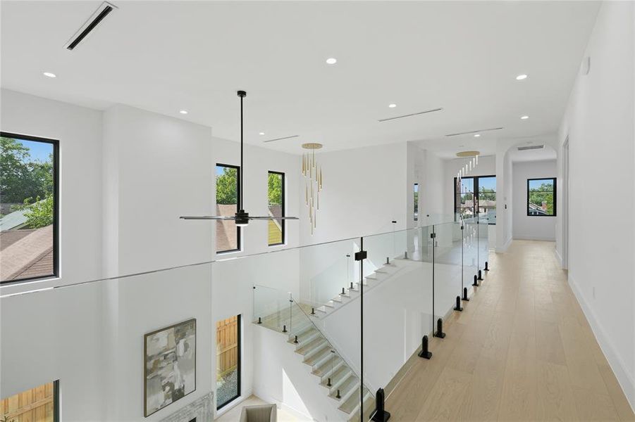 Bright upper-level hallway featuring a glass railing system and light wood-finish flooring Bright upper-level hallway featuring a glass railing system and light wood-finish flooring