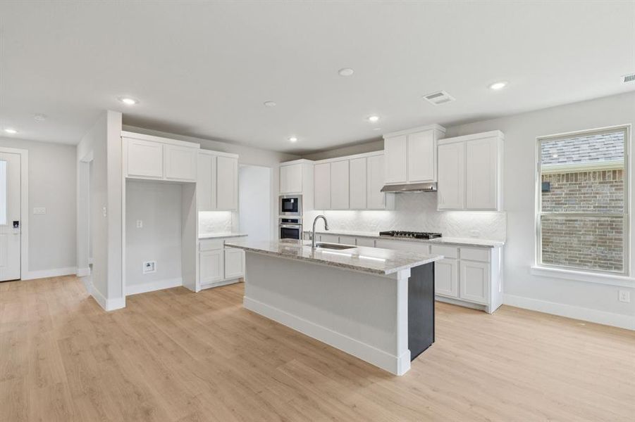 Kitchen with white cabinets, an island with sink, backsplash, light stone countertops, and light wood-style flooring Kitchen with white cabinets, an island with sink, backsplash, light stone countertops, and light wood-style flooring
