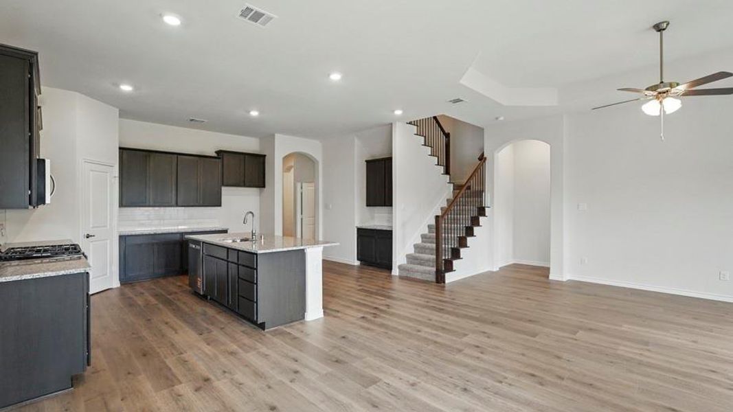 Open-concept living space featuring wood-finish flooring, a contemporary kitchen with dark cabinetry and a central island, a multi-level ceiling with a ceiling fan, a carpeted staircase with an iron spindle railing, and recessed lighting