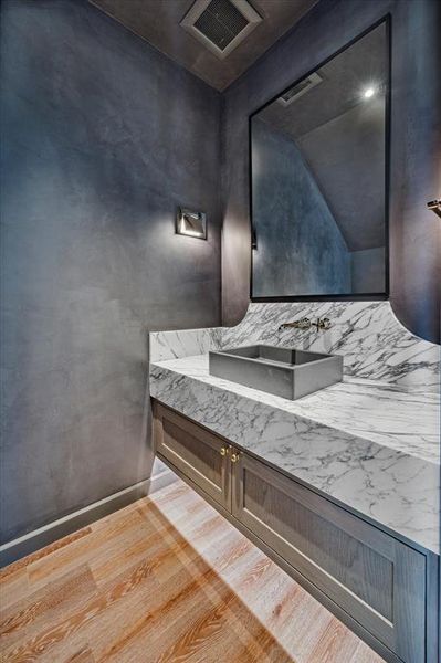 This powder room features sleek, moody lime wash paint walls that create an intimate ambiance. The floating vanity is topped with a striking marble countertop and an integrated backsplash, adding a luxurious touch. A rectangular, vessel-style sink sits atop the counter, paired with a wall-mounted faucet for a streamlined look.