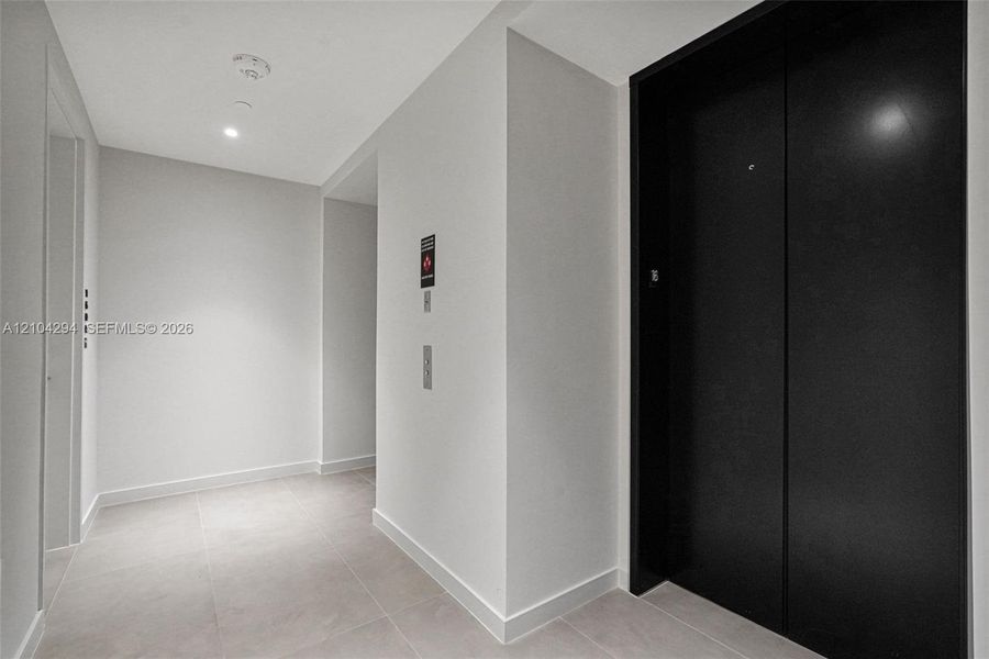 Recessed ceiling lighting illuminates the corridor, featuring a black elevator entrance, light-toned wall finishes, and square tile flooring
