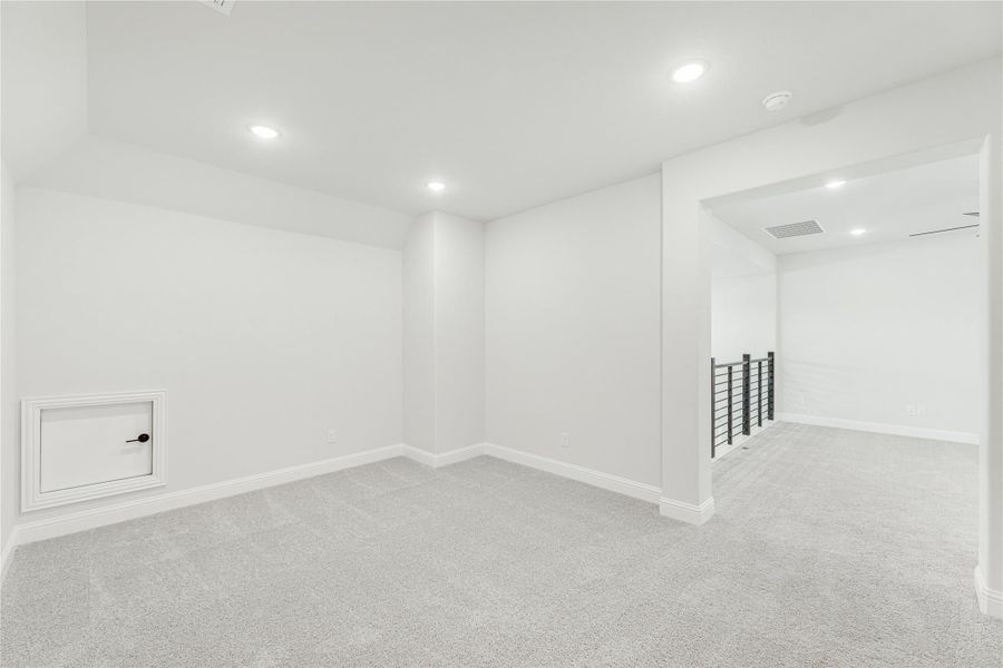 Empty lower-level room with gray carpet, white walls, recessed lighting, and metal stair railing visible in hallway
