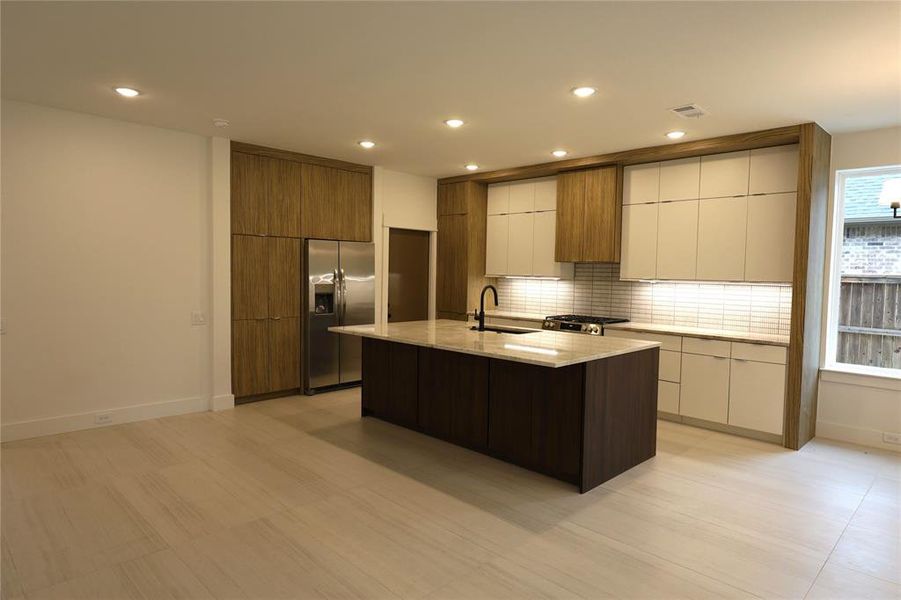 Kitchen with modern cabinets, backsplash, appliances with stainless steel finishes, light stone countertops, and a kitchen island with sink