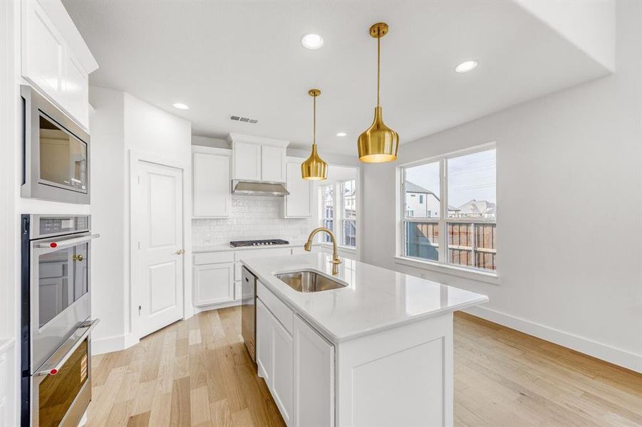 Kitchen with light wood finished floors, an island with sink, white cabinetry, stainless steel appliances, and pendant lighting