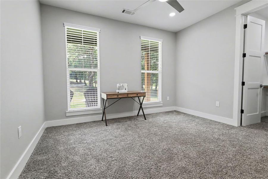 Spacious room featuring neutral carpeting, light gray walls, two windows with blinds, a ceiling fan with integrated lighting, and a white interior door