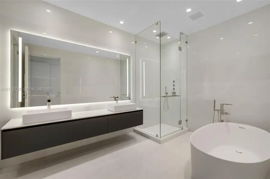 Floating double-vanity with vessel sinks, expansive LED-backlit mirror, glass-enclosed walk-in shower, and freestanding soaking tub