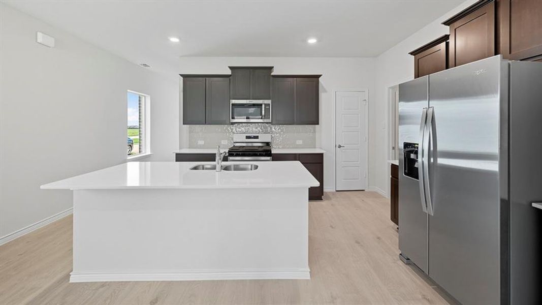 Kitchen with appliances with stainless steel finishes, a center island with sink, backsplash, light wood-style flooring, and light stone counters