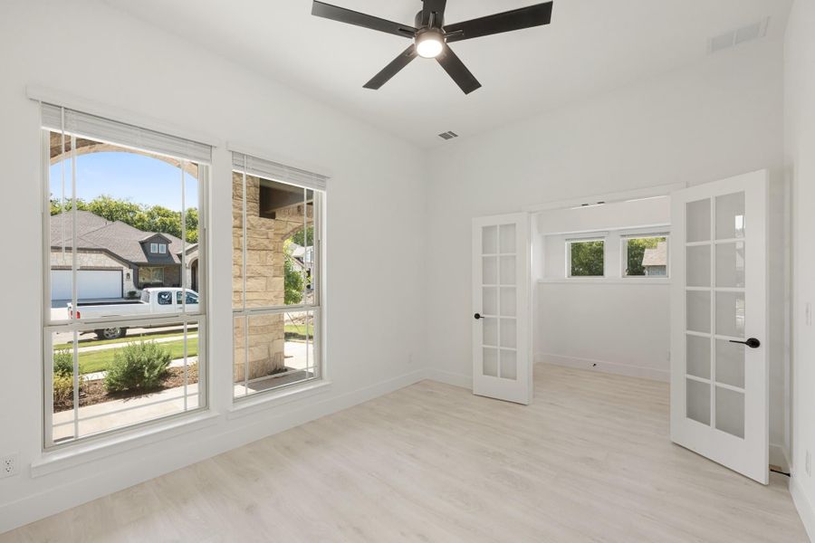 Empty room with light wood floors, ceiling fan, large windows, and white French doors
