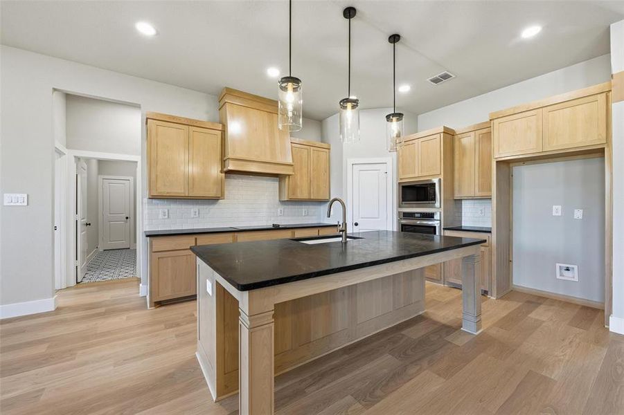 Kitchen featuring light brown cabinetry, tasteful backsplash, hanging light fixtures, a kitchen island with sink, and a kitchen bar