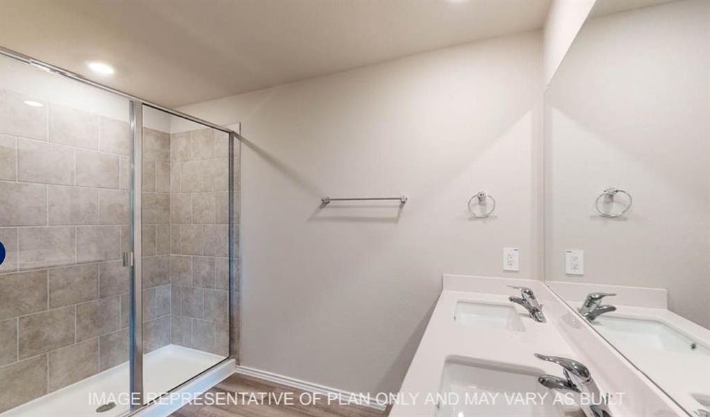 Bathroom featuring a glass-enclosed shower with tile surround, dual undermount sinks set in a white countertop, large wall-mounted mirror, and wood-finish flooring Bathroom featuring a glass-enclosed shower with tile surround, dual undermount sinks set in a white countertop, large wall-mounted mirror, and wood-finish flooring