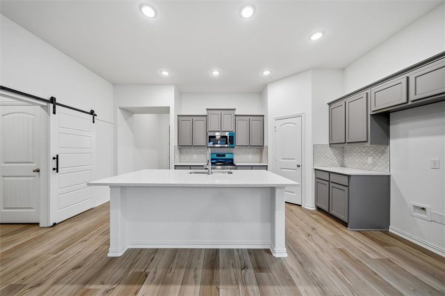 Kitchen with gray cabinetry, a barn door, stainless steel appliances, an island with sink, and recessed lighting