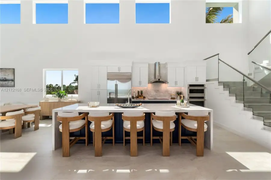 Double-height ceilings with transom windows, large center island with waterfall countertop, stainless steel kitchen appliances, and glass-paneled staircase railing Double-height ceilings with transom windows, large center island with waterfall countertop, stainless steel kitchen appliances, and glass-paneled staircase railing