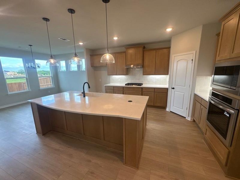 Open-concept kitchen featuring an expansive island with a matte black faucet, wood-finish cabinetry, stainless steel appliances, and wood-finish flooring