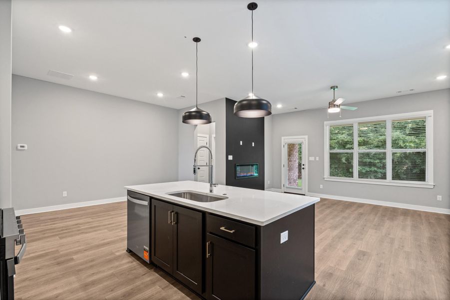 Open-concept living space featuring a central island with an undermount sink and a dishwasher, recessed lighting, wood-finish flooring, a contemporary fireplace, and large windows with blinds Open-concept living space featuring a central island with an undermount sink and a dishwasher, recessed lighting, wood-finish flooring, a contemporary fireplace, and large windows with blinds