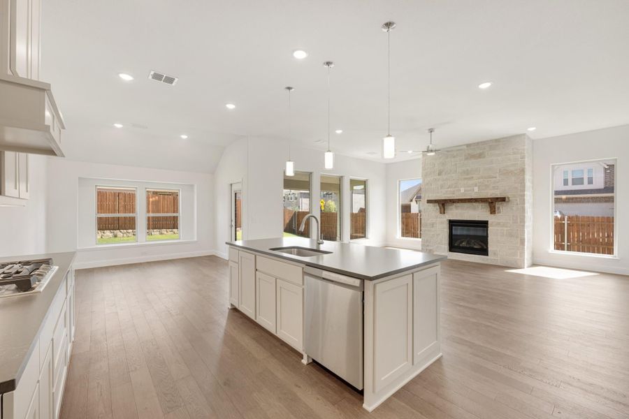 Open-concept kitchen with white island, gray countertops, and stone fireplace in adjoining living area with hardwood floors