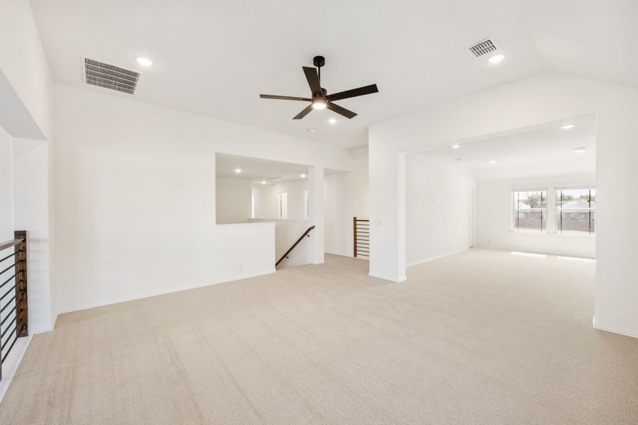 Spacious open loft area with beige carpet, ceiling fan, white walls, and staircase railing