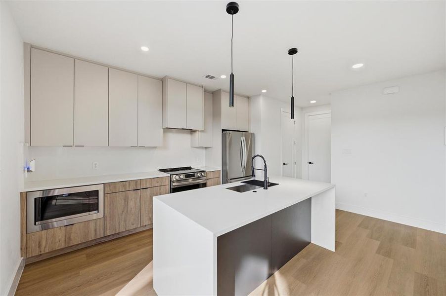 Kitchen features recessed lighting and pendant lighting over the island. Light wood cabinetry extends throughout with built-in microwave. Quartz waterfall island includes a black faucet and undermount sink. Sliding glass door provides access to the backyard patio. Vinyl flooring extends throughout.
