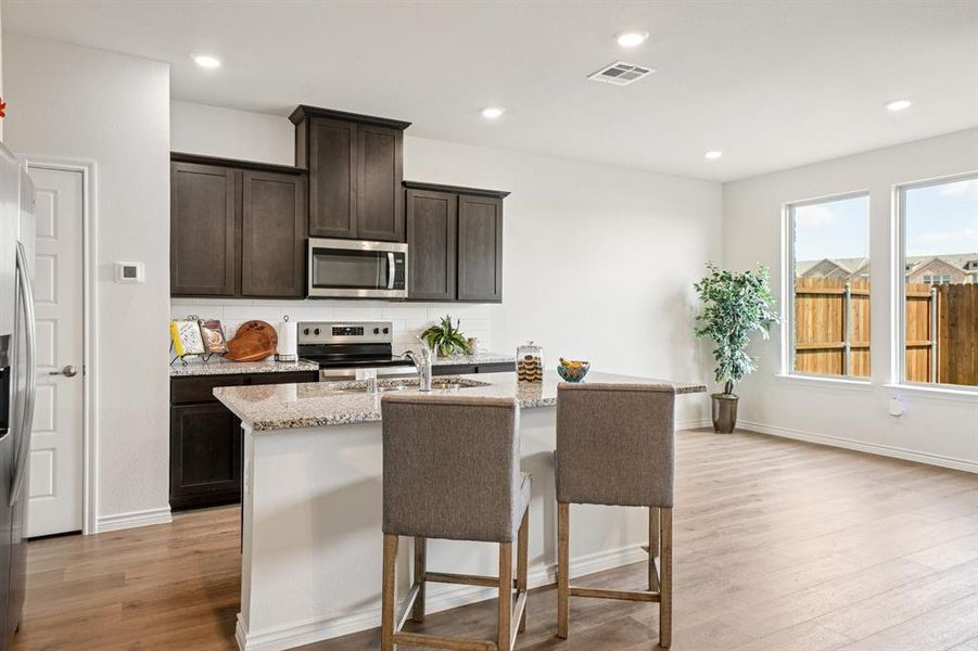 Kitchen with backsplash, dark brown cabinetry, light stone countertops, a center island with sink, and recessed lighting Kitchen with backsplash, dark brown cabinetry, light stone countertops, a center island with sink, and recessed lighting
