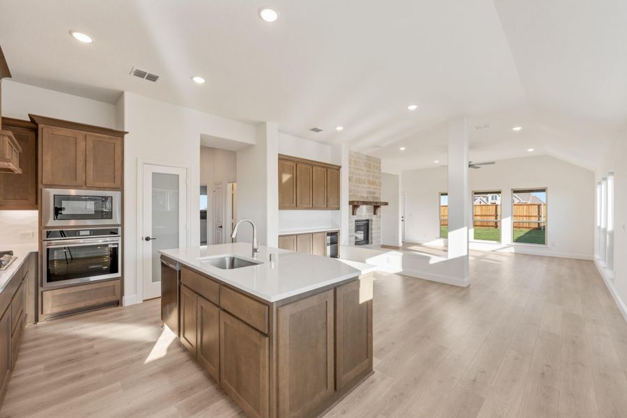 Open-concept kitchen with center island, stainless steel appliances, wood cabinets, and light hardwood floors flowing into living area with fireplace. Open-concept kitchen with center island, stainless steel appliances, wood cabinets, and light hardwood floors flowing into living area with fireplace.