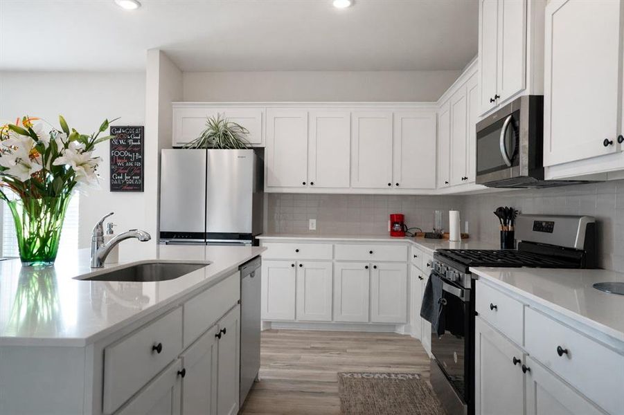 Kitchen with stainless steel appliances, white cabinets, light stone counters, a center island with sink, and light wood-style floors