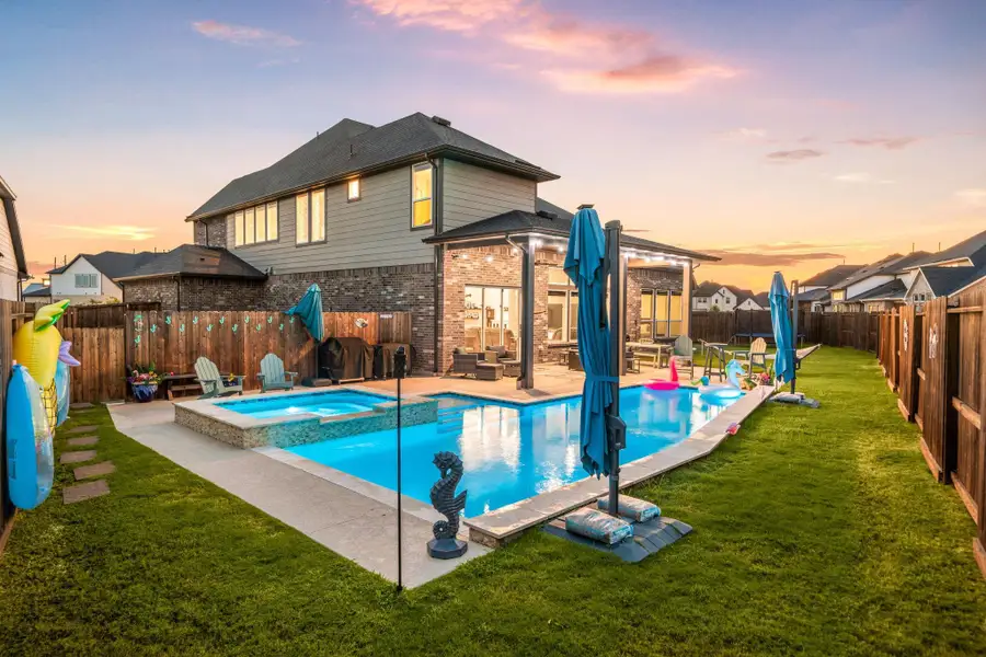 This backyard oasis showcases a sparkling pool and raised spa, framed by a spacious patio and neatly landscaped lawn. The brick exterior and covered patio with recessed lighting create a perfect space for outdoor enjoyment, while the evening sky enhances the inviting atmosphere.