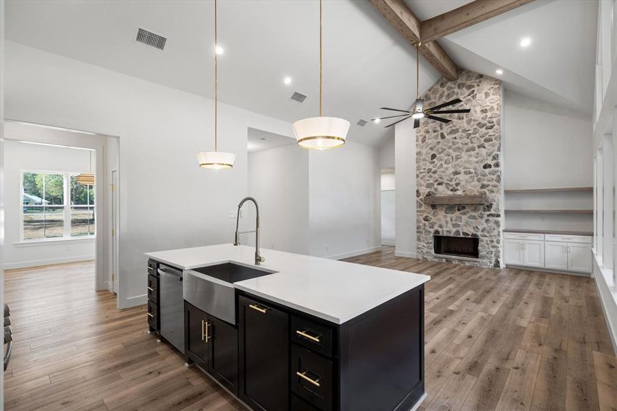 Kitchen featuring dark cabinetry, beam ceiling, pendant lighting, light wood-style flooring, and a stone fireplace