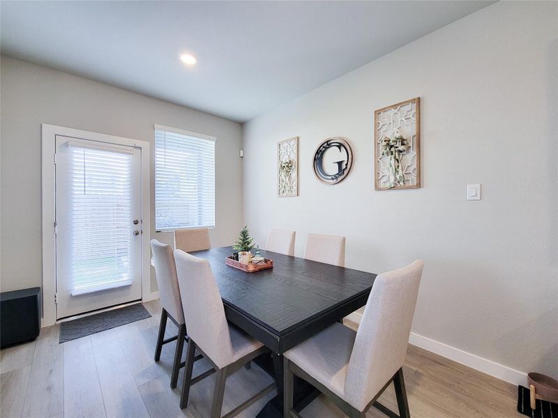 Natural light streams in through a glass door and window with blinds, while decorative wall art adds a touch of elegance. The space features neutral tones and light hardwood flooring, creating a welcoming atmosphere.