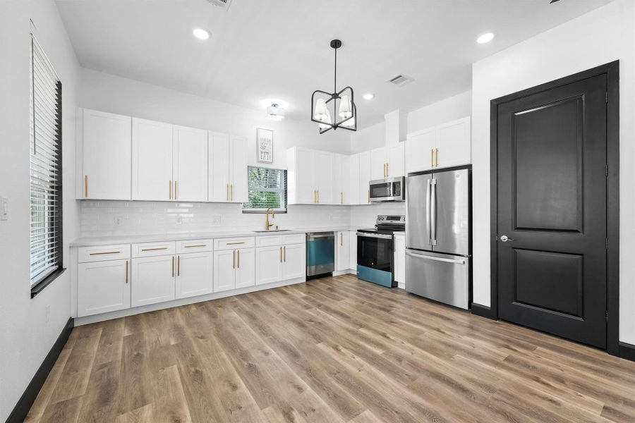 This modern kitchen features sleek white cabinets with gold handles, stainless steel appliances, and a stylish light fixture. It has a spacious layout with wood-look flooring and ample natural light.