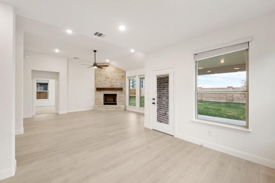 Open living room with stone fireplace, light hardwood floors, ceiling fan, and backyard view through large windows Open living room with stone fireplace, light hardwood floors, ceiling fan, and backyard view through large windows