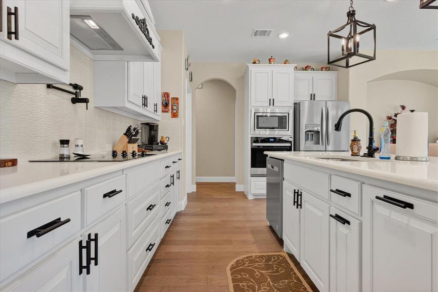 Kitchen with white cabinetry, arched walkways, custom range hood, stainless steel appliances, and decorative light fixtures Kitchen with white cabinetry, arched walkways, custom range hood, stainless steel appliances, and decorative light fixtures
