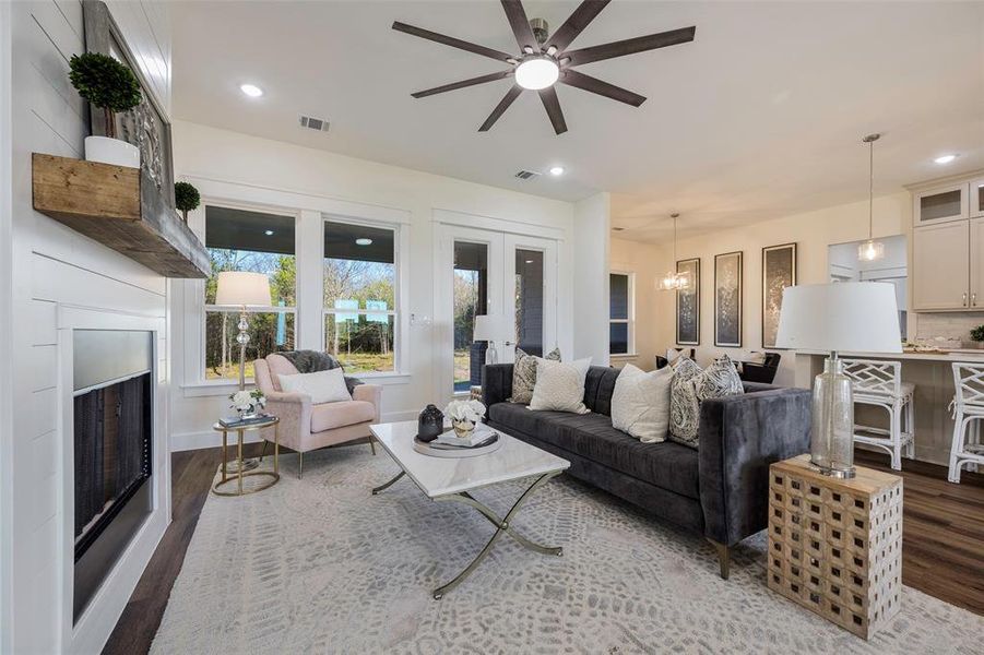 Inviting living room with striking shiplap fireplace wall and rustic mantel creates the perfect gathering space. Expansive windows and glass doors flood the room with natural light and showcase peaceful views. Open concept design flows seamlessly into gourmet kitchen, while rich hardwood flooring anchors the sophisticated modern farmhouse aesthetic. Glass double-doors lead out to covered back porch that overlook the tranquil trees.