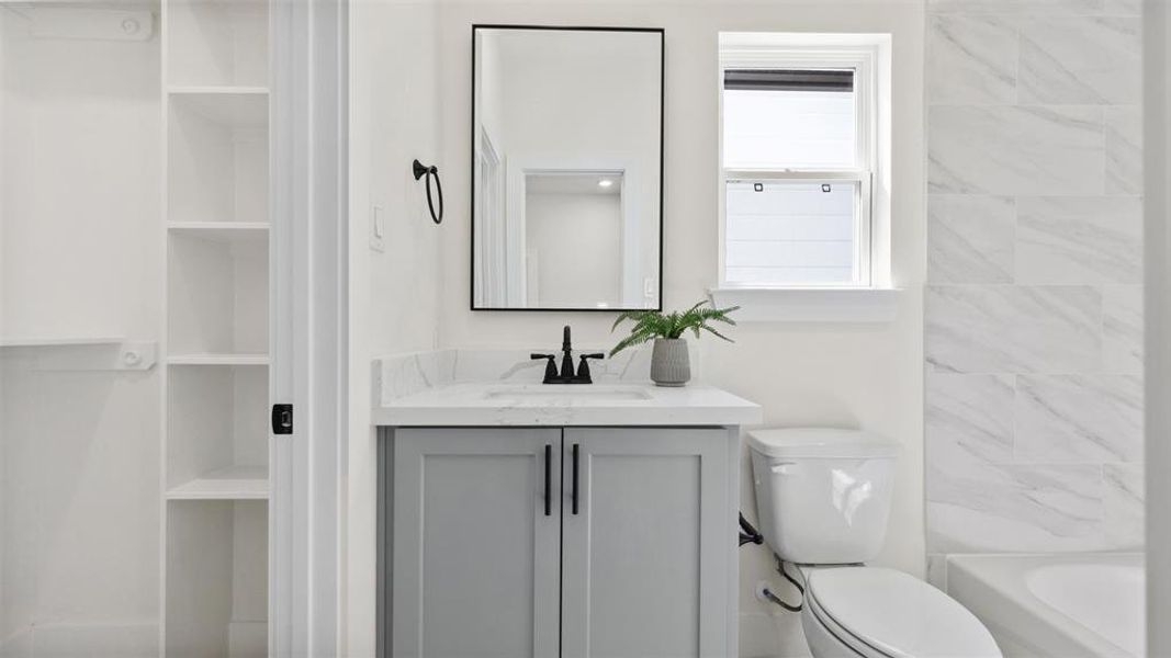 This bathroom features a modern design with a stylish vanity, black fixtures, and a large mirror. It has a bright, airy feel with natural light from a window, built-in shelving, and elegant marble-like wall tiles in the shower area.