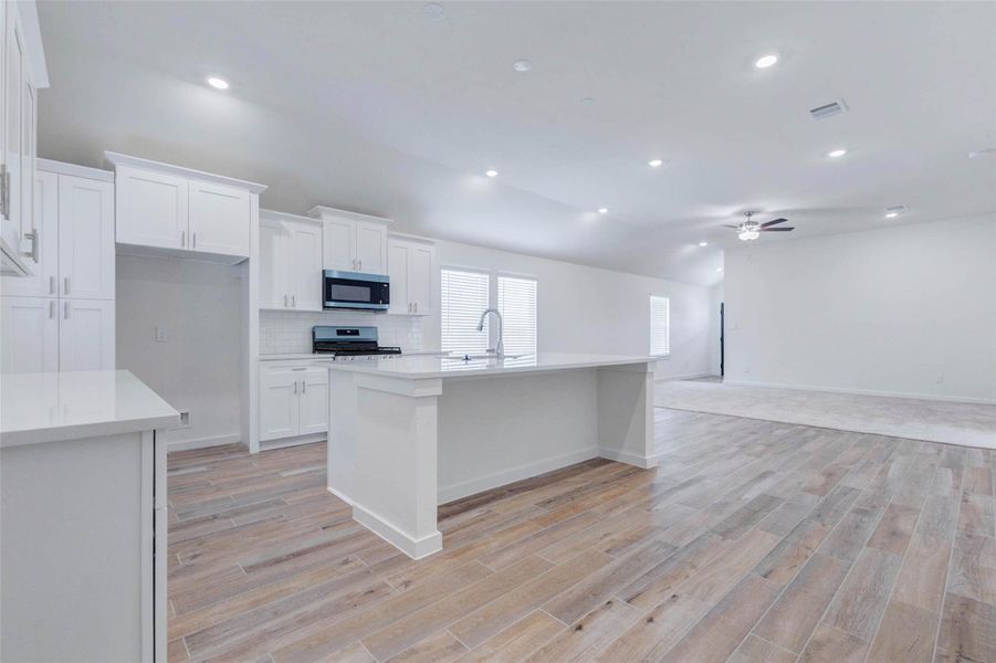 Expansive open-concept view highlighting the kitchen, dining area, and family room in one bright, connected space. White shaker cabinetry, a large center island, sleek countertops, stainless appliances, recessed lighting, wood-look tile flooring, and high ceilings create a fresh modern feel, while the seamless layout keeps everyday living and entertaining easy.